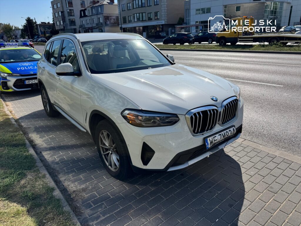 Biały samochód marki BMW zaparkowany na chodniku obok radiowozu policji w miejskim otoczeniu.