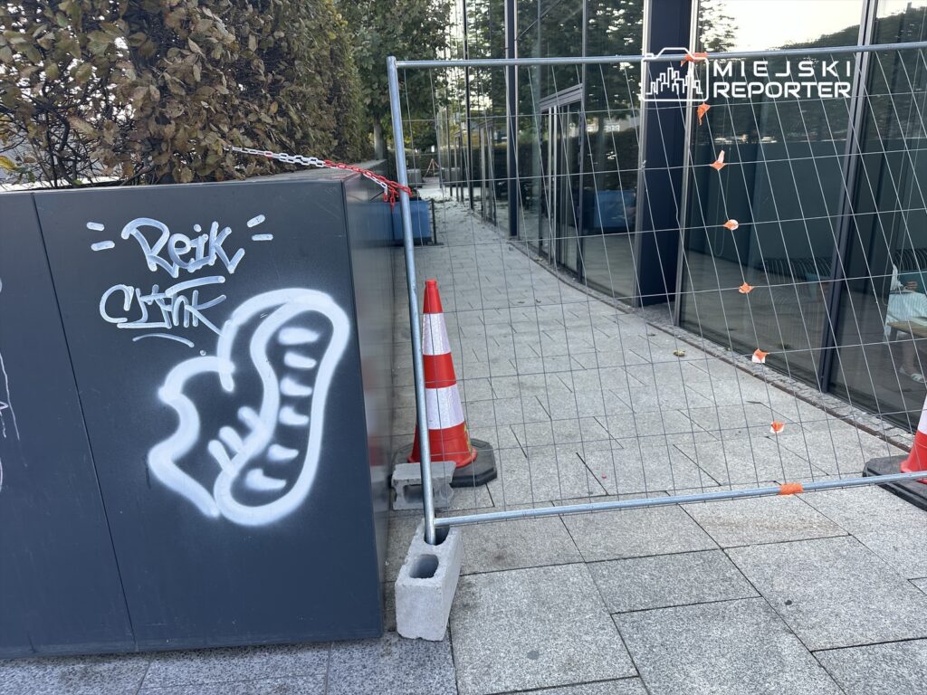 Na szarej ścianie widoczny jest graffiti z napisem „Reik” oraz rysunkiem buta, obok metalowe ogrodzenie z pomarańczowymi pachołkami.