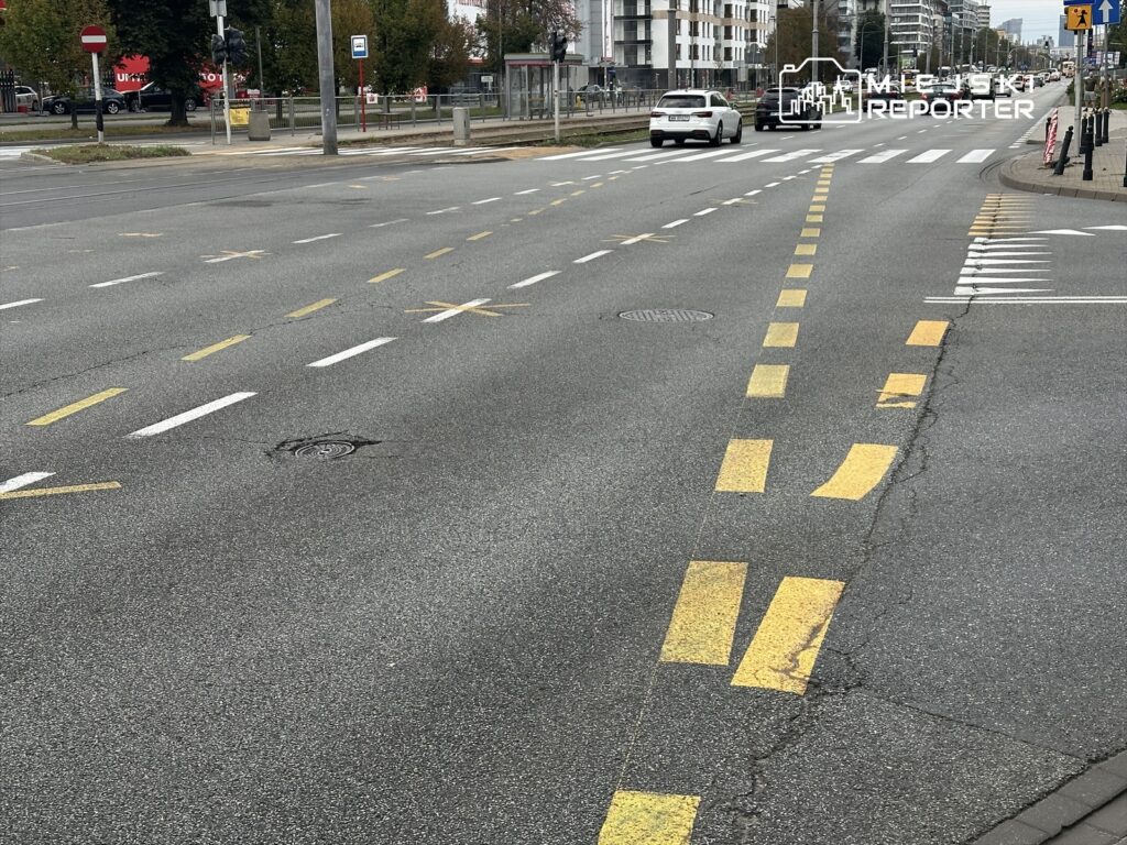 Pusta jezdnia z wyraźnymi liniami drogowymi w żółtym kolorze, w tle widoczne budynki mieszkalne i sygnalizacja świetlna.