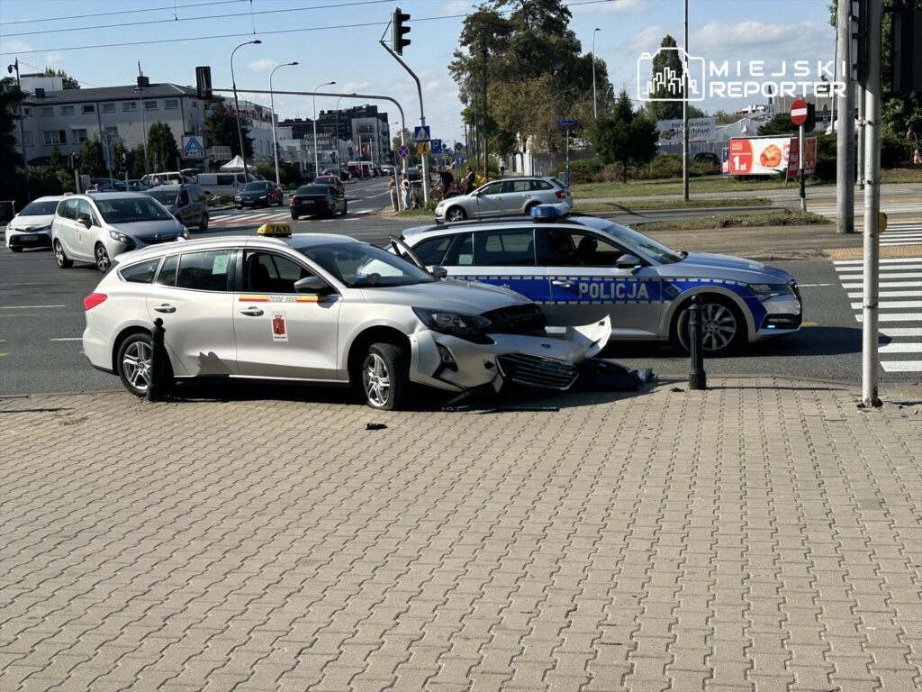 Zderzenie dwóch samochodów na skrzyżowaniu, w tym taksówki z widocznym uszkodzeniem przodu oraz radiowozu policyjnego.