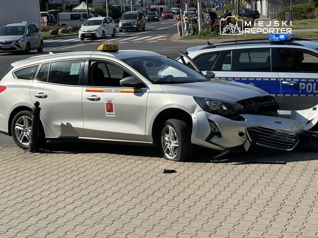 Zderzenie taksówki z policyjnym radiowozem na ruchliwej ulicy, uszkodzona przednia część pojazdów.