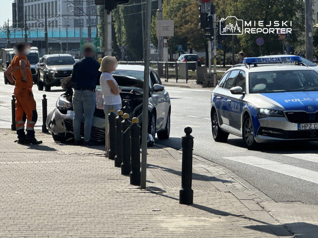 Mężczyzna w niebieskiej koszuli rozmawia z kobietą w białej bluzce obok uszkodzonego samochodu, a w tle stoi radiowóz policji.