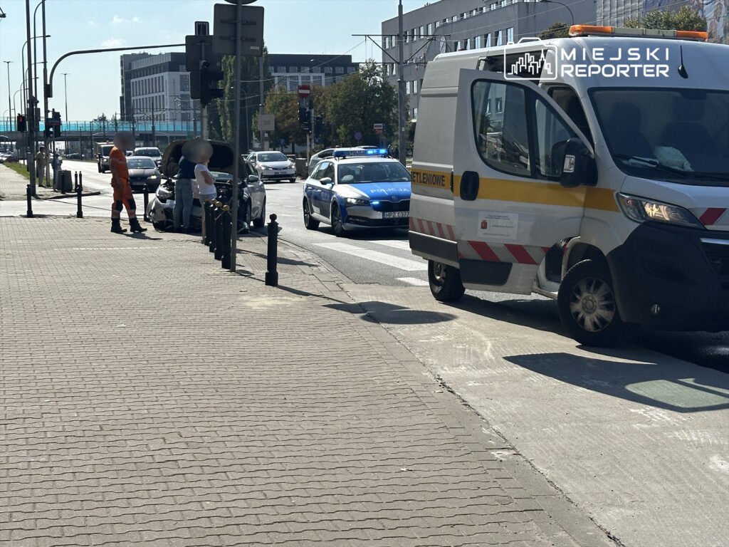 Mężczyzna w pomarańczowym uniformie pomaga kierowcy z uniesioną maską samochodu na ulicy, obok stoi radiowóz policji.