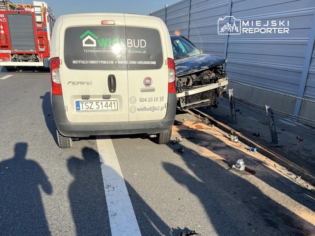 Zniszczony samochód osobowy leży na poboczu drogi, obok stoi biały furgon z logo firmy budowlanej.