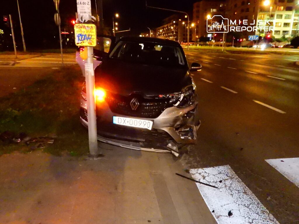 Uszkodzony samochód osobowy marki Renault stoi na chodniku, zderzony z słupem drogowym, w nocy na ruchliwej ulicy.