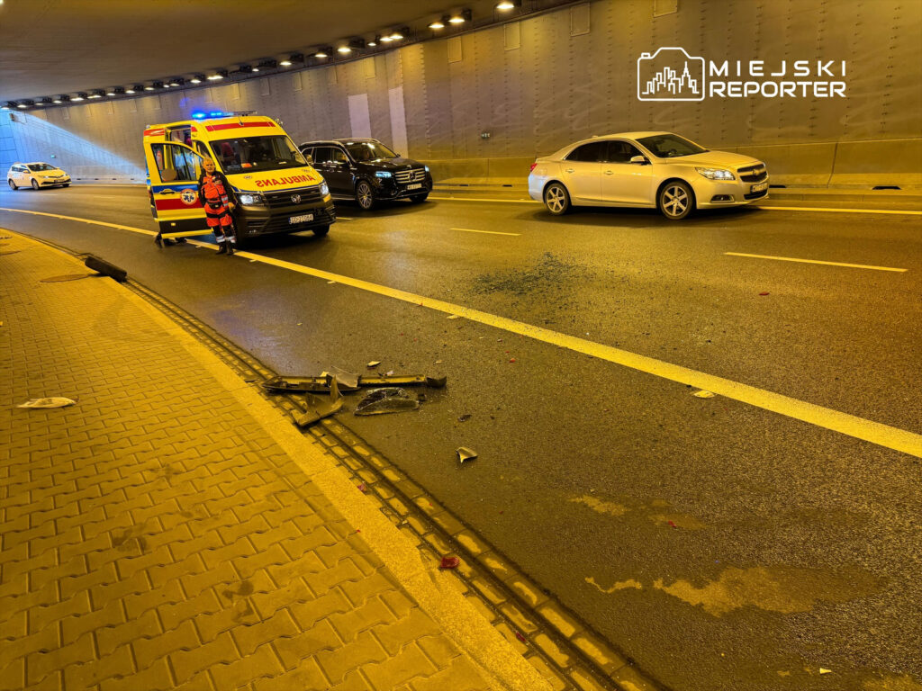 Karetka z włączonymi sygnałami stoi w tunelu, obok leżą fragmenty rozbitego pojazdu na jezdni.