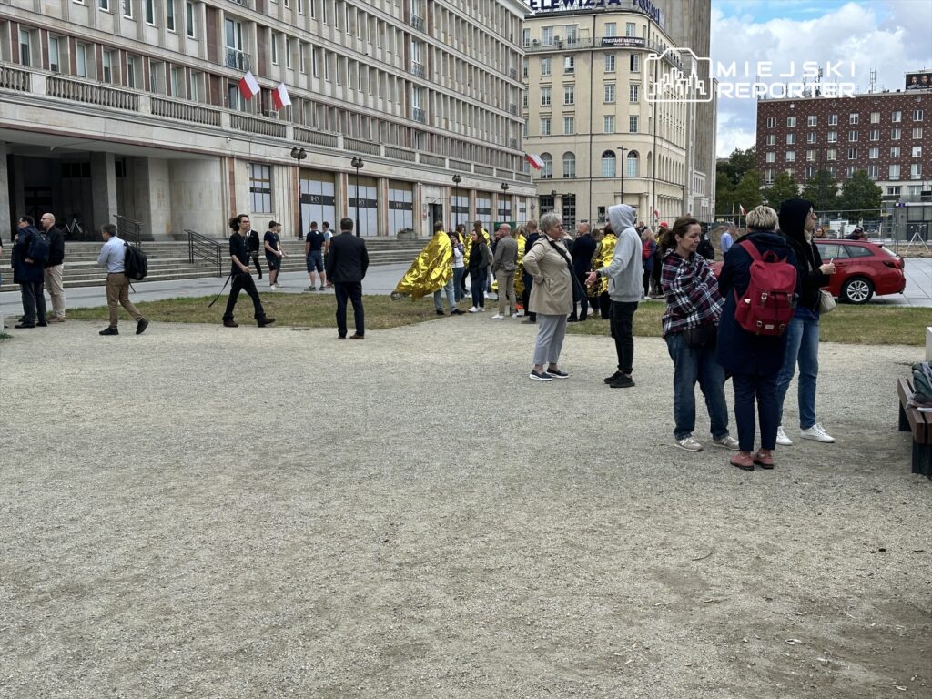 Grupa osób rozmawia na placu przed nowoczesnym budynkiem, niektórzy trzymają transparenty i wstążki.