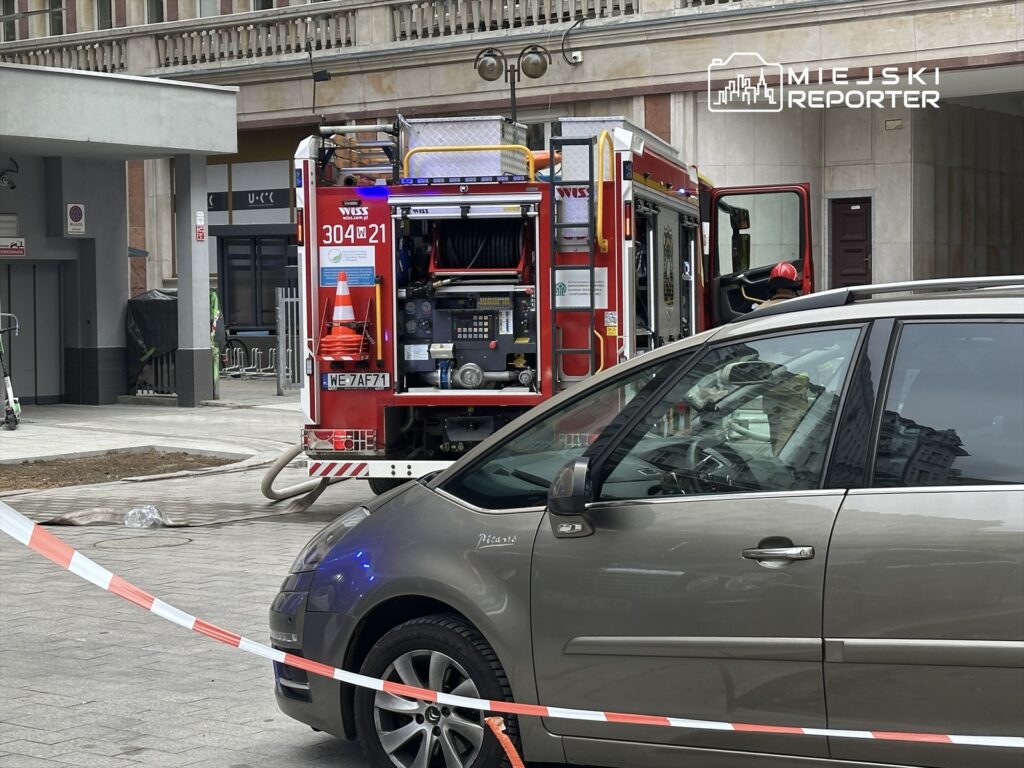 Straż pożarna z włączonymi sygnałami świetlnymi stoi na ulicy, obok zaparkowanego samochodu, w otoczeniu budynków.