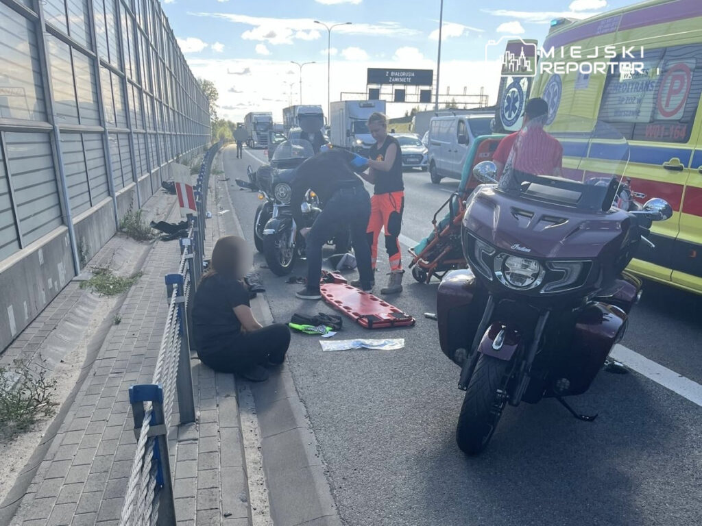 Grupa osób udziela pomocy poszkodowanemu motocykliście na ruchliwej drodze, obok karetki pogotowia.