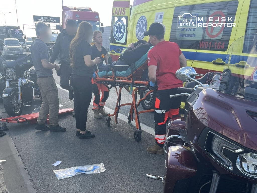 Grupa ratowników medycznych przenosi poszkodowanego na noszach w pobliżu zablokowanej drogi, obok motocykla.