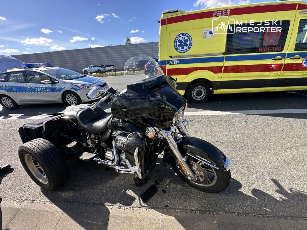 Czarny motocykl z uszkodzoną tylną częścią stoi na poboczu drogi, obok radiowozu policji i karetki pogotowia.