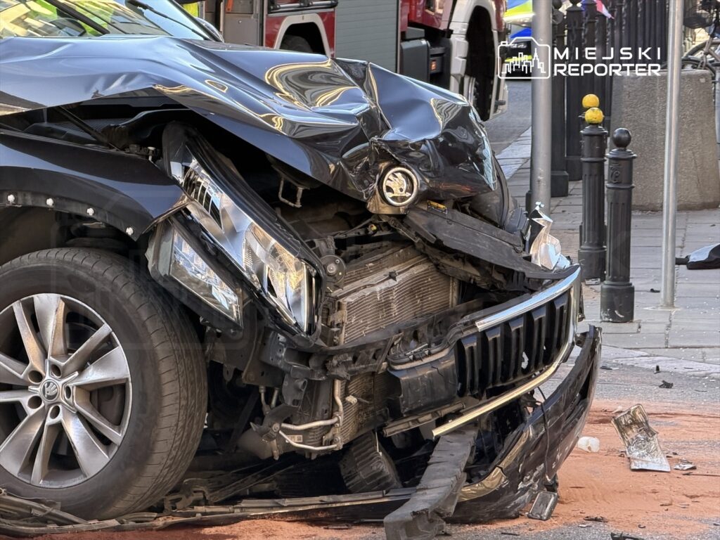 Zniszczony czarny samochód osobowy z wgniecionym przodem stoi na ulicy, otoczony fragmentami wraku i piaskiem.