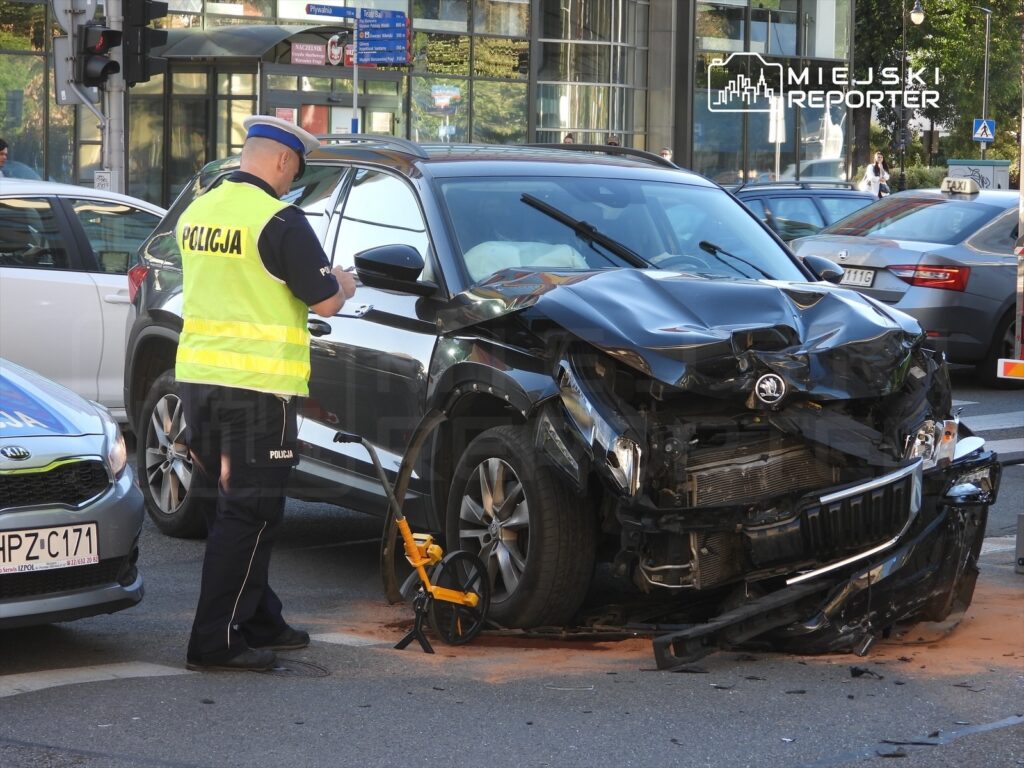Policjant w kamizelce odblaskowej sprawdza zniszczony czarny samochód osobowy po wypadku drogowym na skrzyżowaniu.