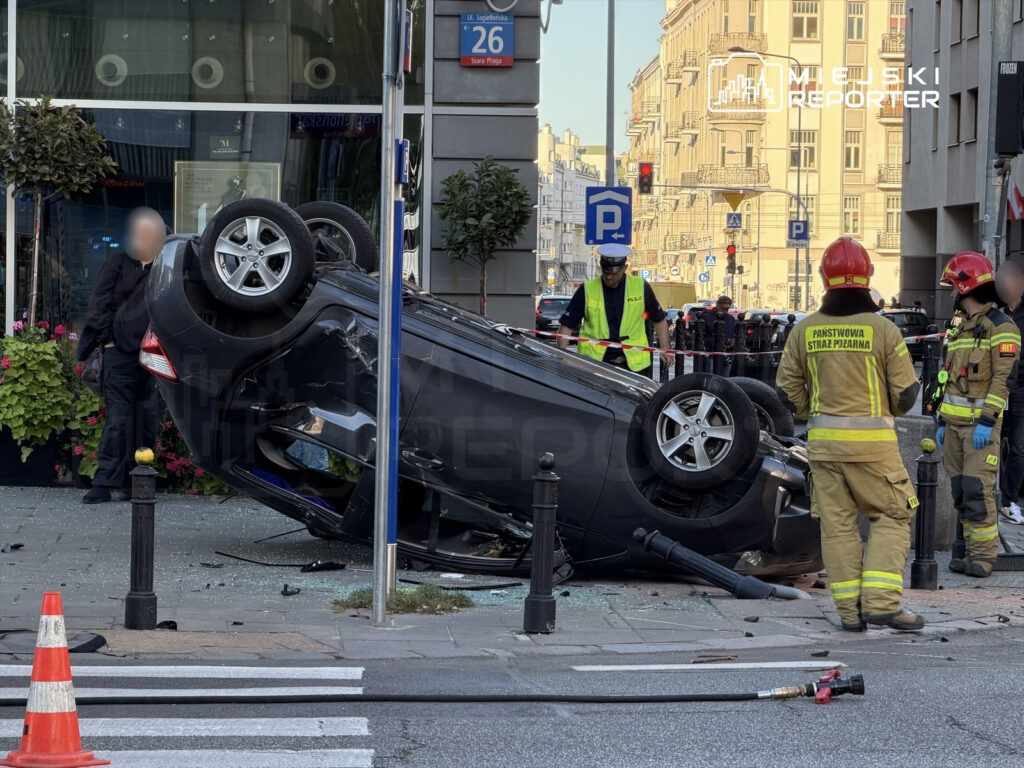 Czarny samochód leży na dachu obok budynku, a strażacy i policjanci zabezpieczają miejsce zdarzenia.