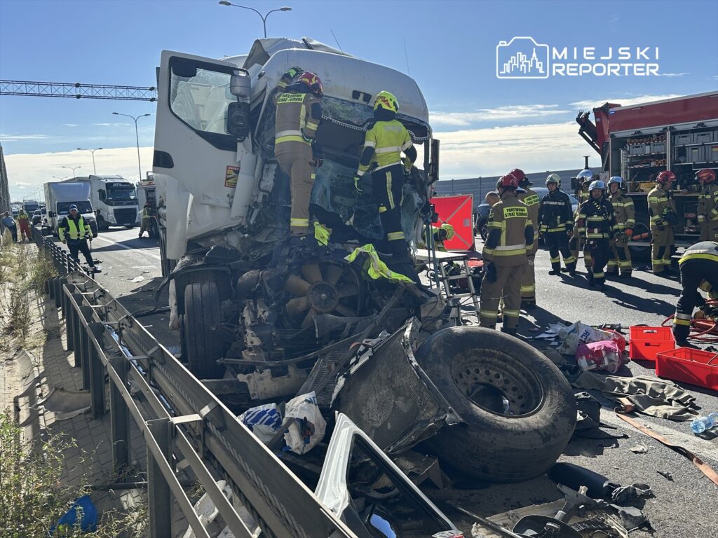 Grupa strażaków pracuje przy rozbitej ciężarówce na autostradzie, obok stoją inne pojazdy.