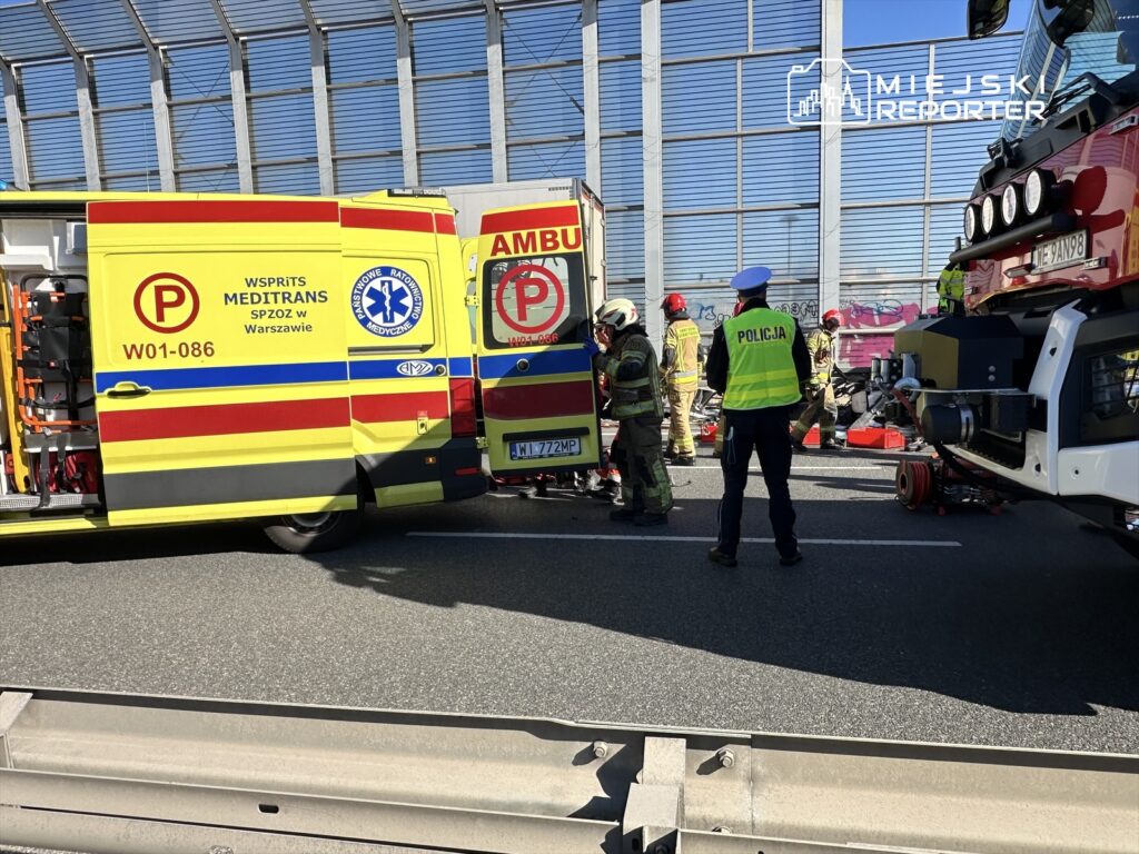 Ratownicy w mundurach pracują przy wypadku drogowym na autostradzie, obok wozów strażackich i ambulansu.