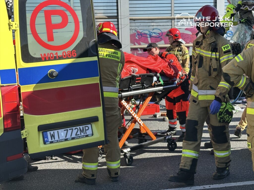 Ratownicy medyczni i strażacy udzielają pomocy poszkodowanemu na noszach w pobliżu karetki.
