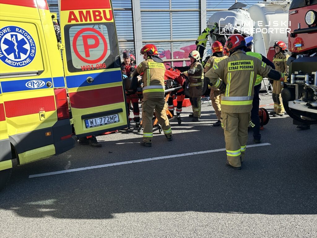 Grupa ratowników medycznych i strażaków udziela pomocy poszkodowanemu na drodze, obok dwóch ambulansów.