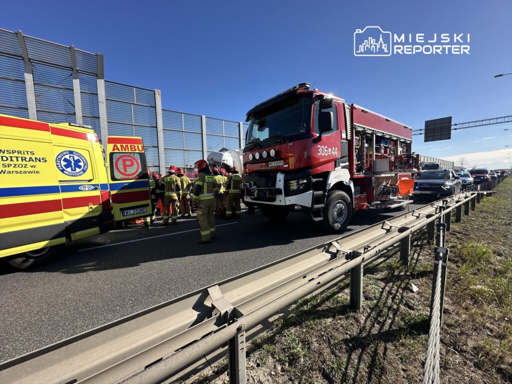 Zespół ratunkowy w mundurach pracuje przy wypadku drogowym na autostradzie, obok wozu strażackiego i karetki.