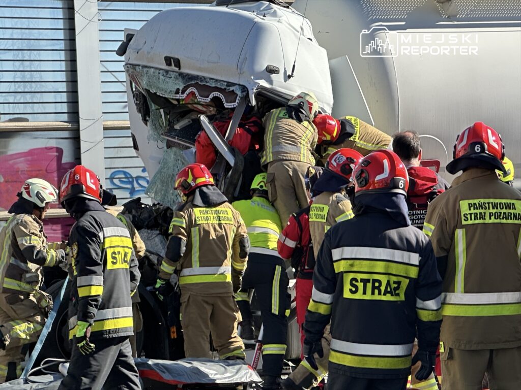 Grupa strażaków w mundurach i hełmach pracuje przy wypadku drogowym, usuwając zniszczony pojazd.