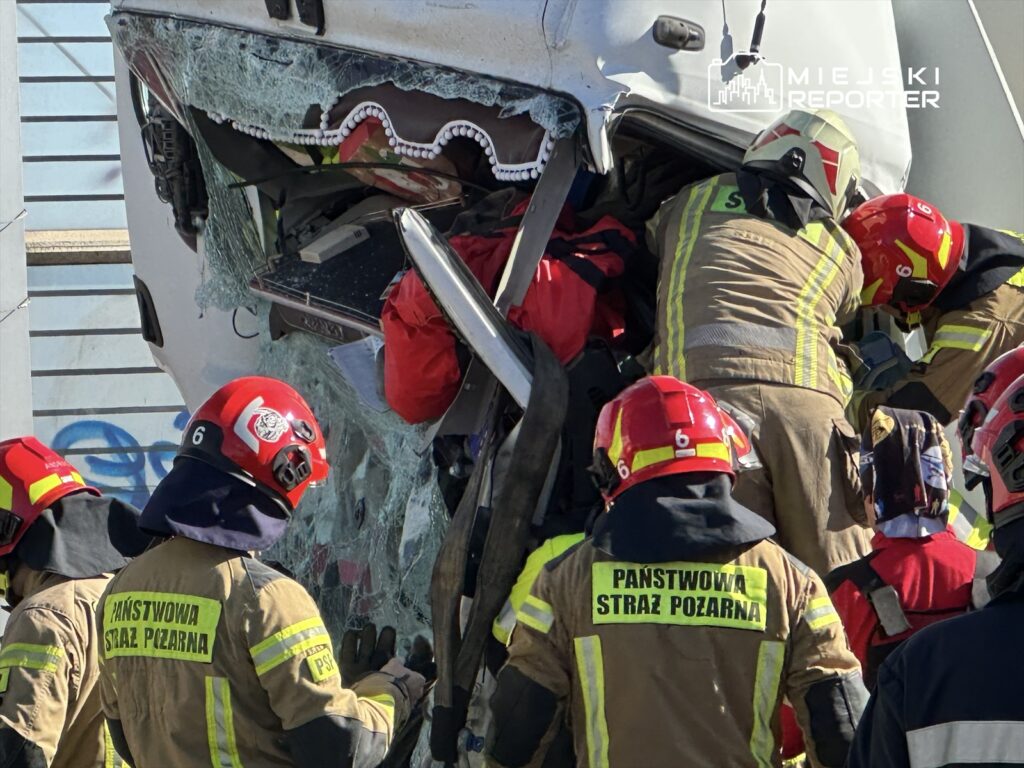Strażacy w mundurach i hełmach pracują nad wydobyciem osoby z rozbitego pojazdu, otoczeni przez zniszczoną kabinę.