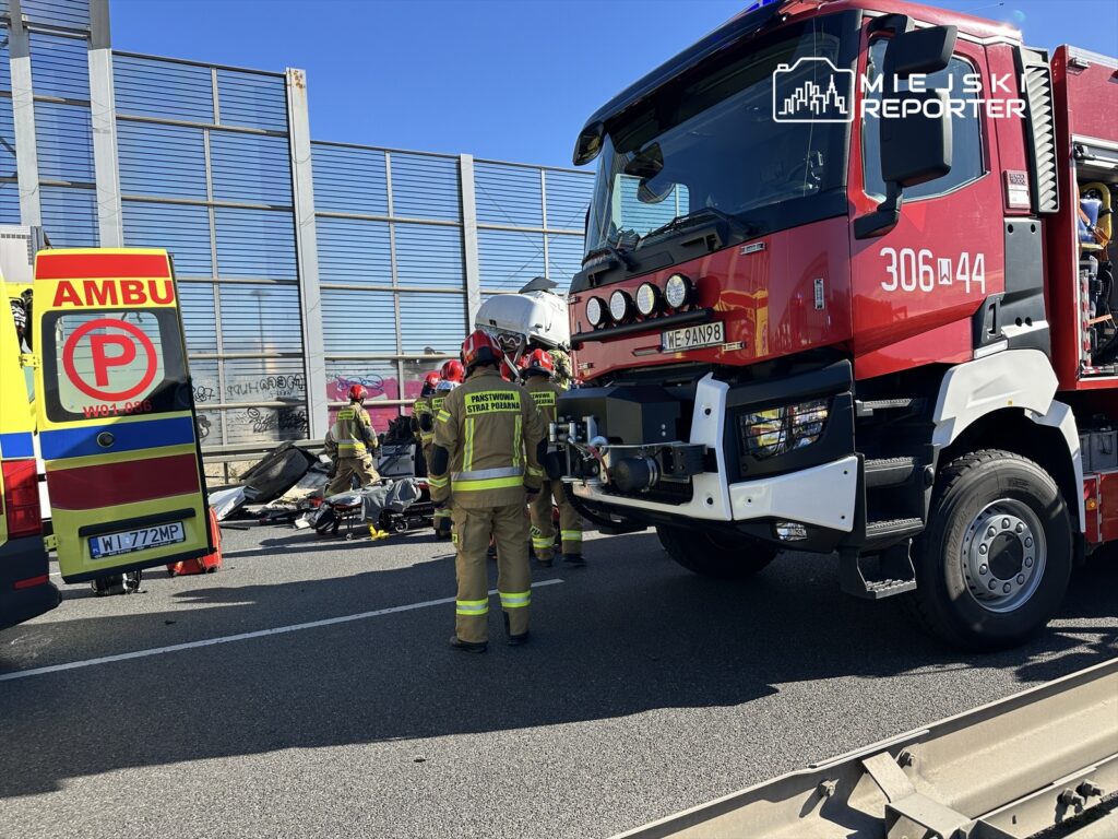 Ratownicy w hełmach pracują przy wypadku drogowym na autostradzie, obok wozu strażackiego i karetki.