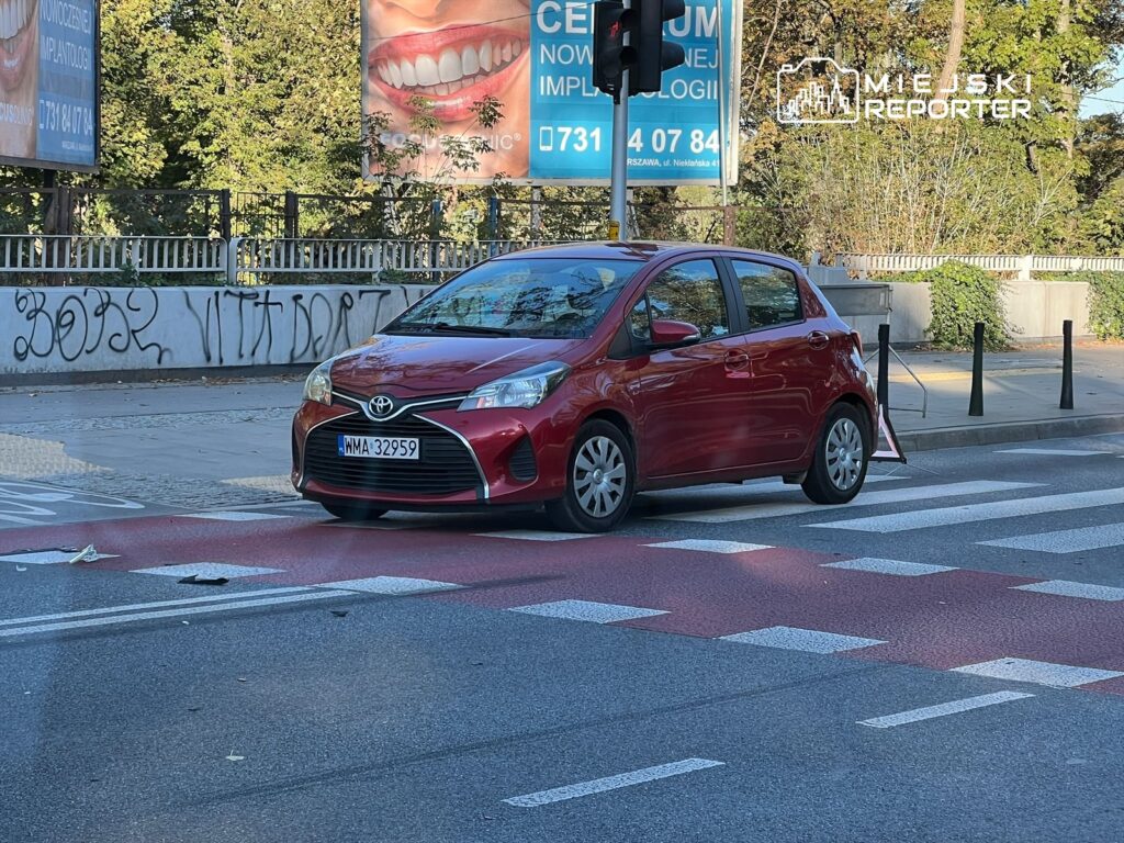 Czerwony samochód osobowy zatrzymany na pasach w miejskim otoczeniu, w tle billboardy reklamowe.