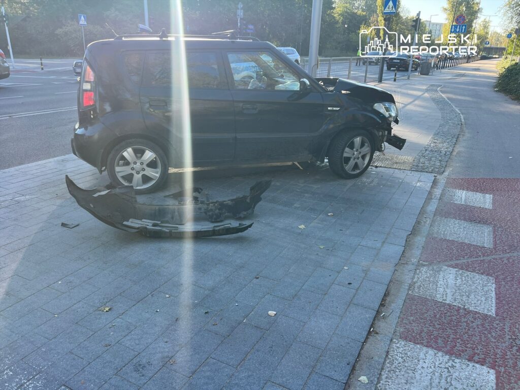 Czarny samochód osobowy z uszkodzonym przodem stoi na chodniku, obok leży zdemontowany zderzak.