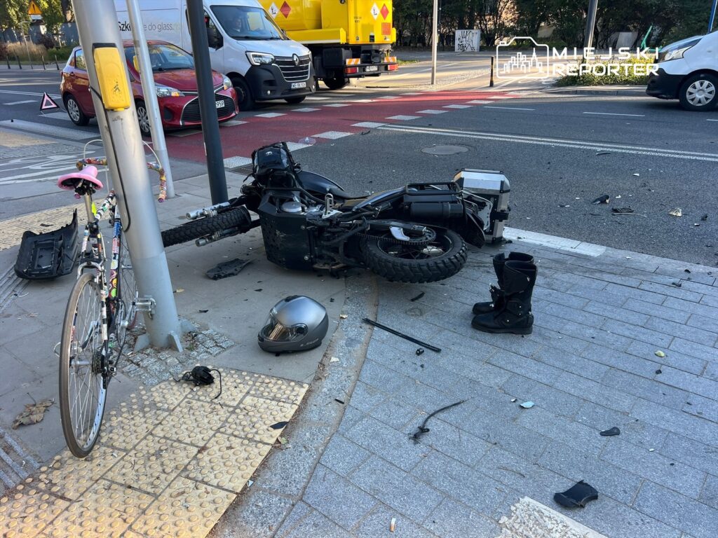 Motocykl leży na boku obok latarni, a obok niego znajduje się rower oraz kask. W tle widać samochody na ulicy.