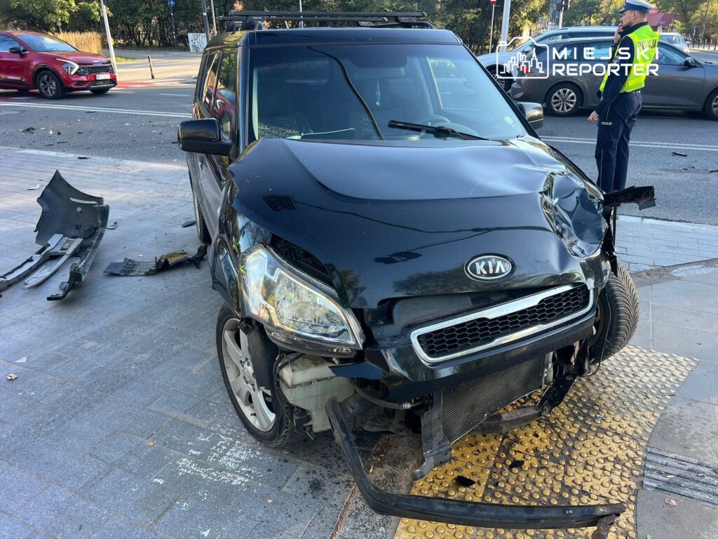 Czarny samochód osobowy z widocznymi uszkodzeniami stoi na chodniku, obok policjant rozmawia przez telefon.