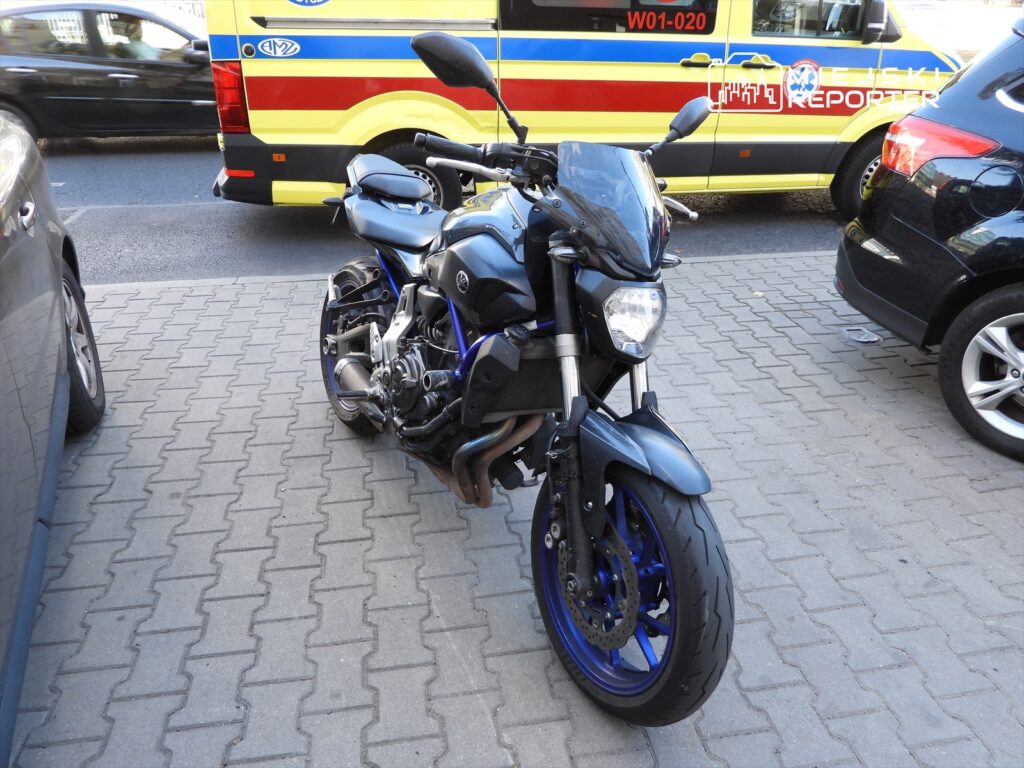 Motocykl marki Yamaha zaparkowany na chodniku obok zaparkowanych samochodów. W tle widoczny pojazd służbowy.