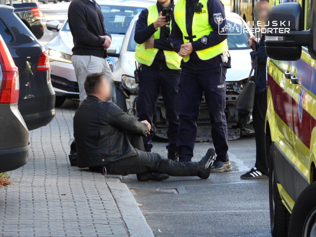 Mężczyzna w skórzanej kurtce siedzi na chodniku, otoczony przez policjantów na miejskiej ulicy.