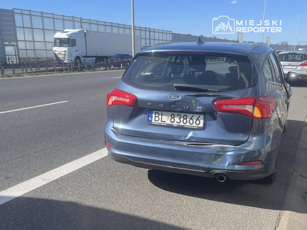 Uszkodzony niebieski samochód marki Ford Focus zaparkowany na poboczu drogi, obok ruchliwej jezdni z ciężarówkami.