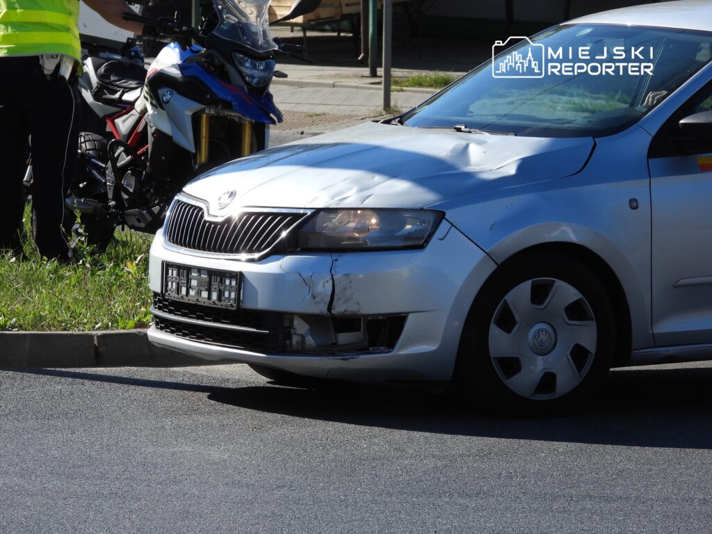 Uszkodzony samochód osobowy zaparkowany przy drodze, obok stoi policjant w odblaskowej kamizelce.