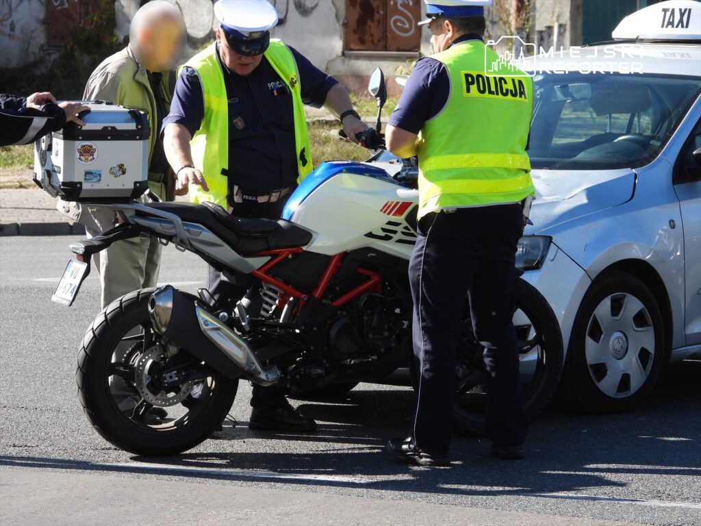 Policjant w kamizelce odblaskowej rozmawia z kierowcą, obok stoi motocykl policyjny i uszkodzone auto.