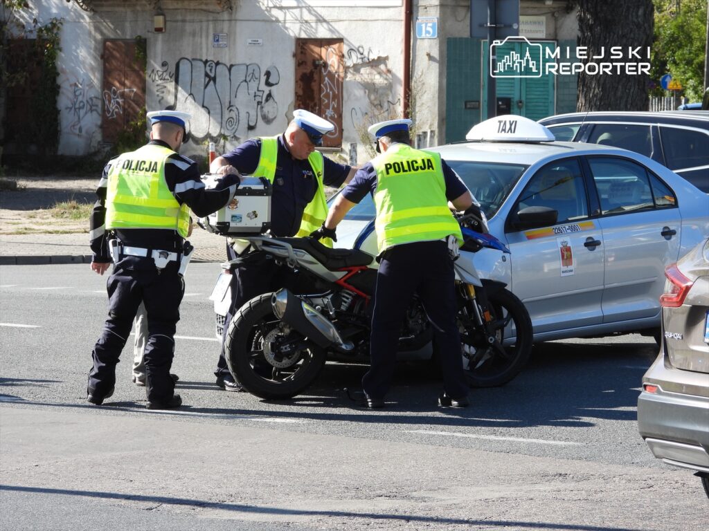 Dwaj policjanci w kamizelkach odblaskowych sprawdzają motor na ulicy, obok zatrzymanego taksówki.