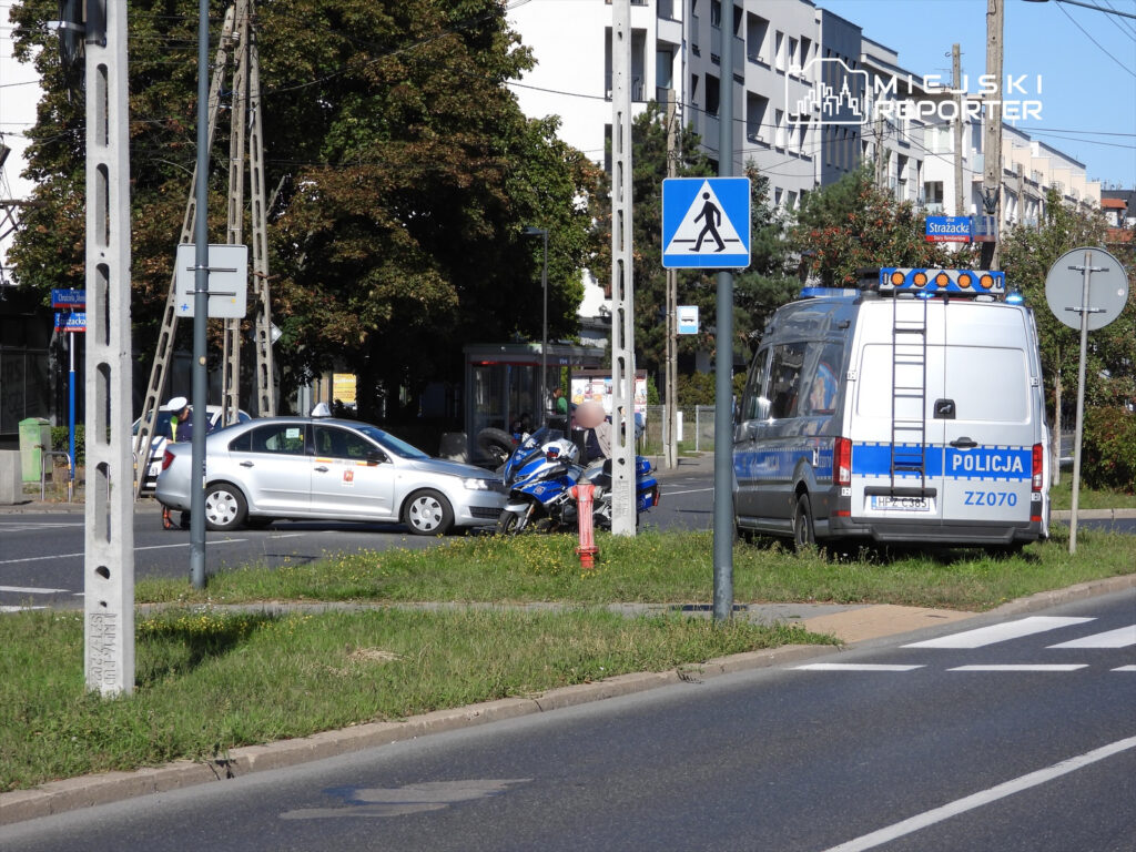 W zdarzeniu drogowym uczestniczy srebrny samochód osobowy oraz policyjny van z włączonymi sygnałami świetlnymi.