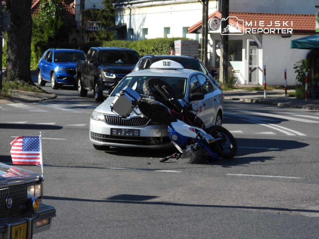 Motocykl leży na jezdni obok taksówki, a w tle widać inne pojazdy na ruchliwej ulicy.