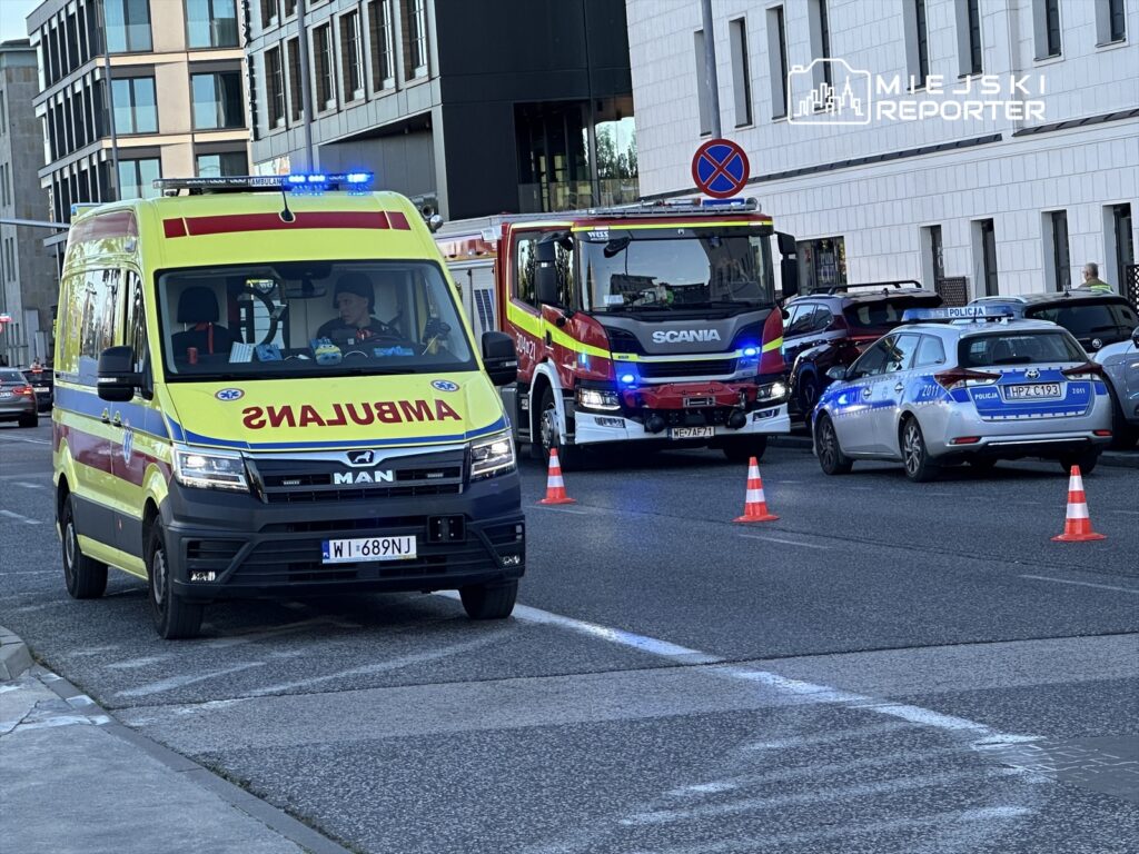 Karetka pogotowia z włączonymi sygnałami świetlnymi zatrzymana na ulicy obok wozu strażackiego i radiowozu policji.