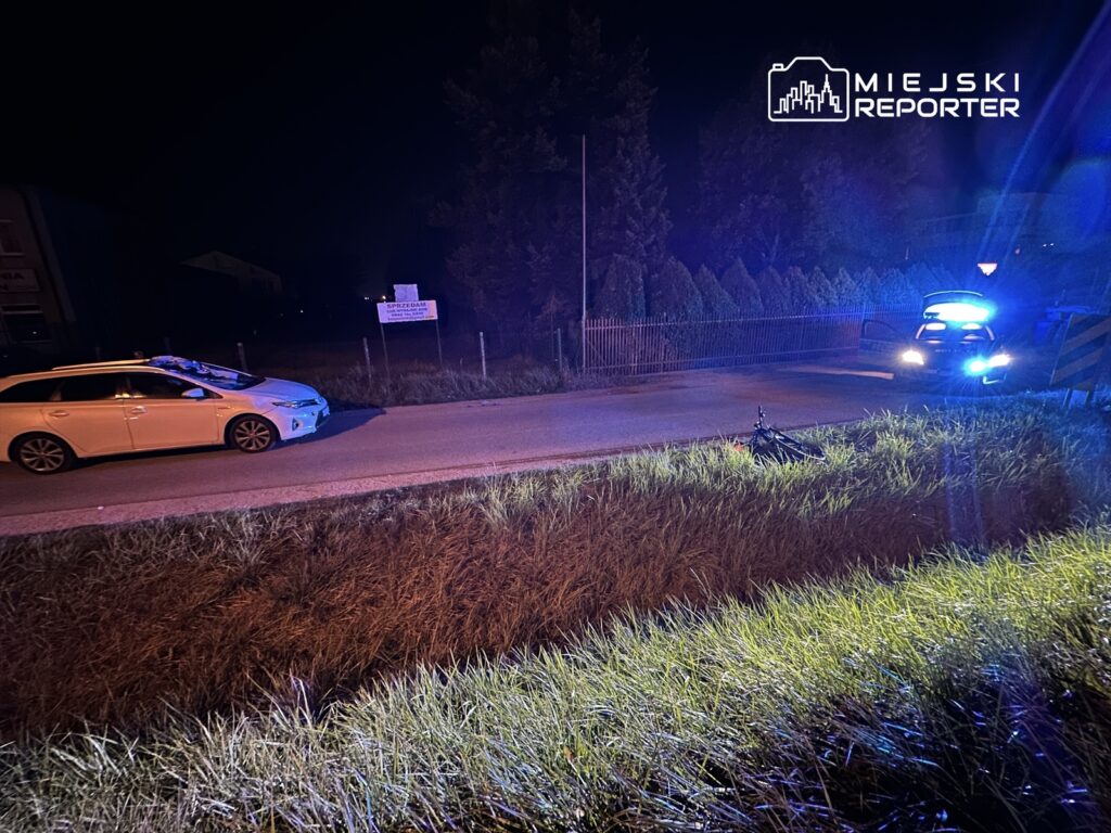 Na drodze stoi biały samochód osobowy obok nieoznakowanego radiowozu policyjnego z włączonymi sygnałami świetlnymi. W tle widać ogrodzenie i znak drogowy. W trawie leży rower.