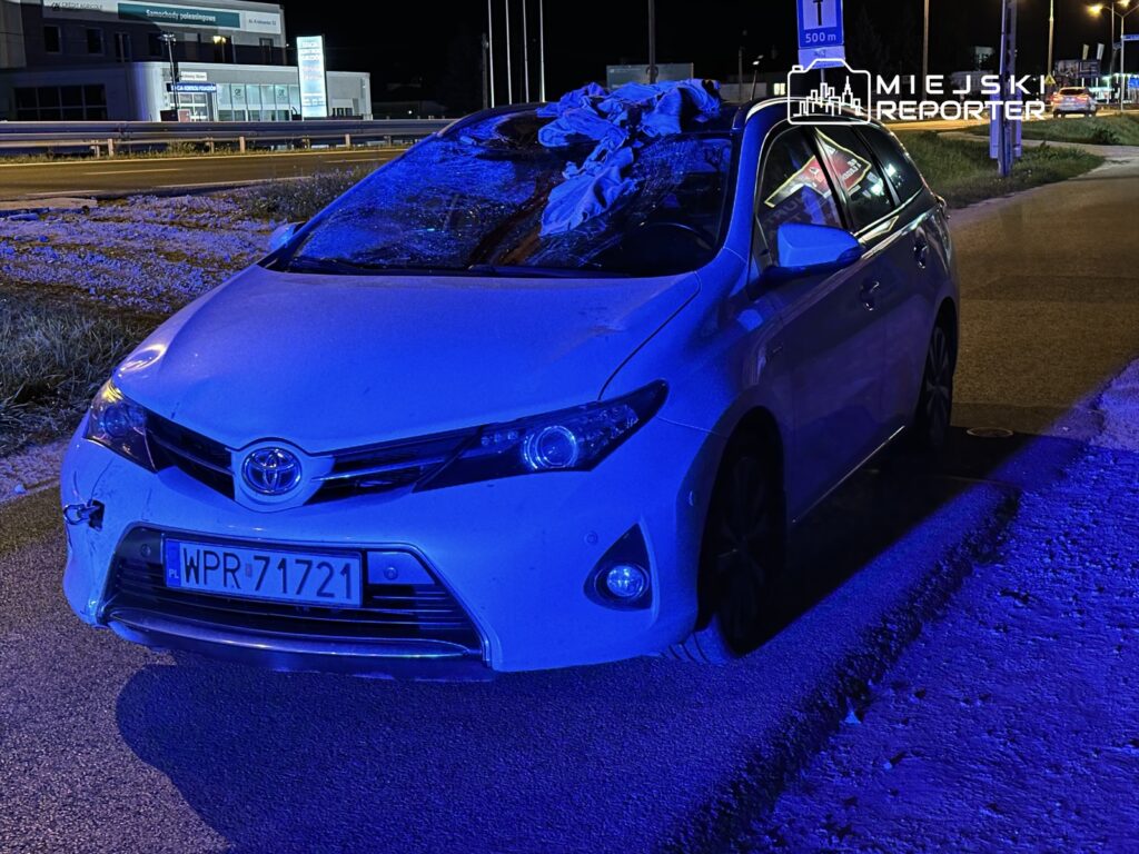 Uszkodzony biały samochód osobowy marki Toyota zaparkowany na poboczu drogi, oświetlony niebieskimi światłami.