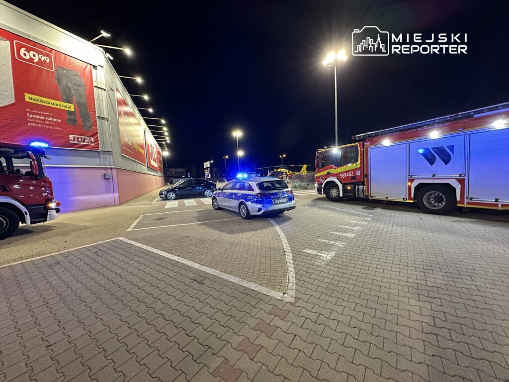 W nocy na parkingu przed dużym sklepem z widocznymi reklamami, straż pożarna i policyjny radiowóz interweniują.