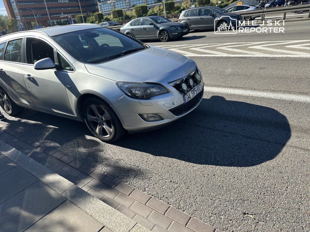 Srebrny samochód osobowy zaparkowany na ulicy, z widocznymi dodatkowymi światłami na przednim zderzaku.