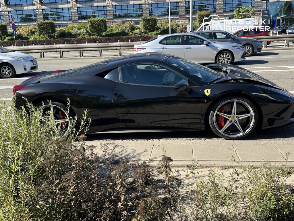 Czarny samochód sportowy marki Ferrari zaparkowany obok ruchliwej ulicy, w tle widać inne pojazdy i nowoczesne budynki.