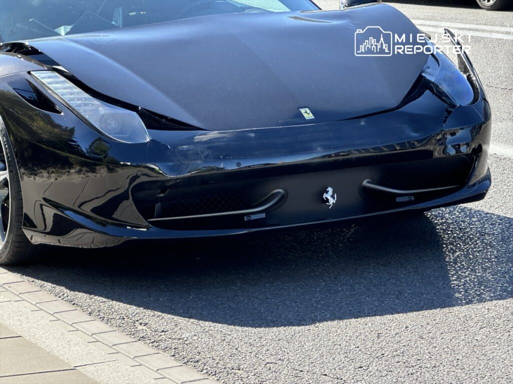 Czarny samochód sportowy z uszkodzoną maską zaparkowany na ulicy, w otoczeniu asfaltu i chodnika.