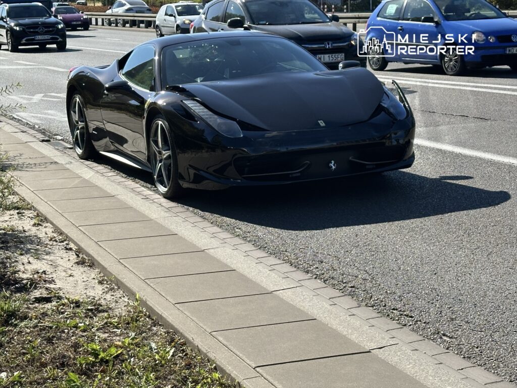 Czarny sportowy samochód Ferrari zaparkowany na chodniku obok ruchliwej drogi, w tle inne pojazdy.