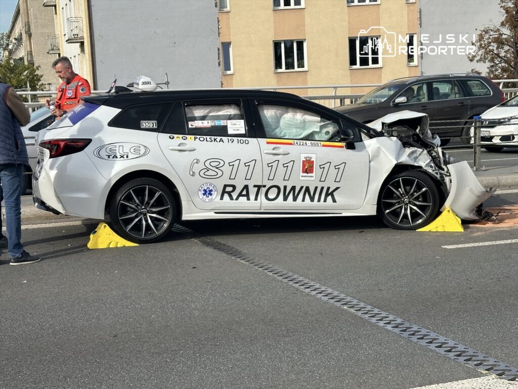 Biały samochód taxi z uszkodzeniem przodu stoi na ulicy, obok niego mężczyzna w czerwonym uniformie rozmawia przez telefon.