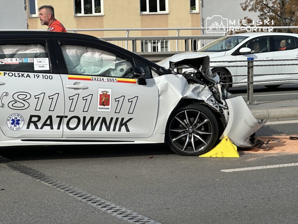 Ratownik w czerwonym uniformie przemieszcza się obok uszkodzonego samochodu na ulicy, w tle widoczne inne pojazdy.