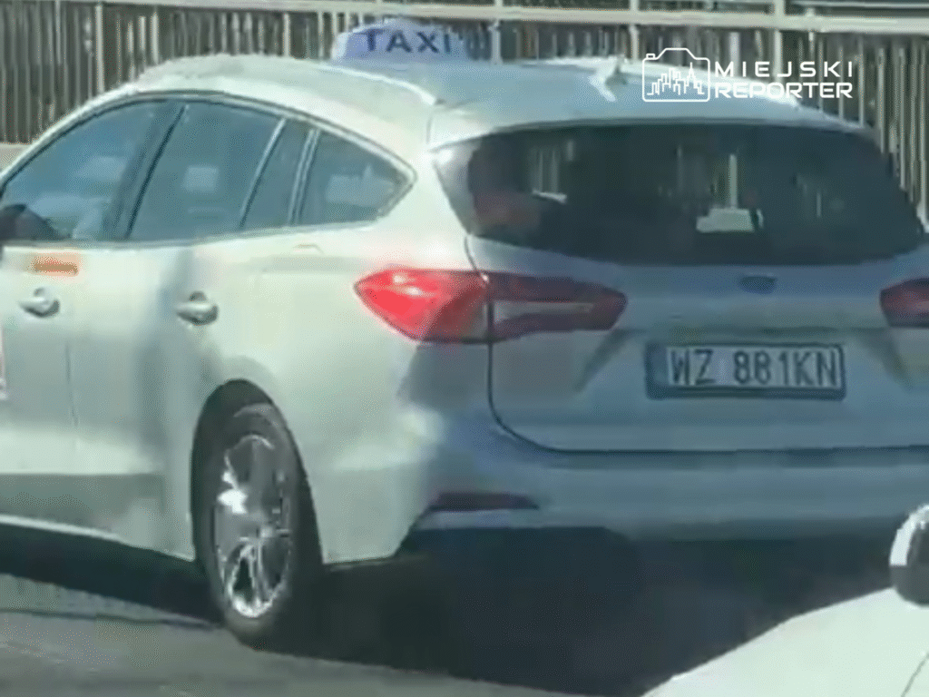 Białe taxi z oznaczeniem na dachu porusza się po drodze, z widoczną tablicą rejestracyjną WZ 881KN.