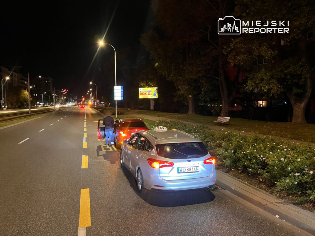 Mężczyzna w ciemnej kurtce wsiada do samochodu osobowego na oświetlonej ulicy nocą, obok stoi taksówka.